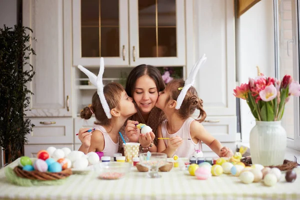 Two little sisters with white rabbits ears on their heads  kiss their mother dyeing the eggs for the Easter table in the cozy light kitchen