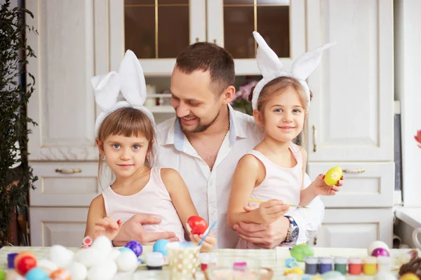 Boldog apa és a két kis lánya a fejükön fehér nyúl füle festék a tojás a húsvét tábla a hangulatos fény konyhában — Stock Fotó