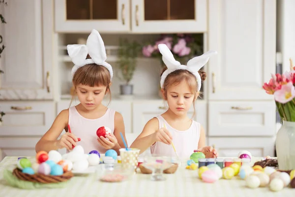 Zwei kleine Schwestern mit weißen Hasenohren auf dem Kopf färben die Eier für den Ostertisch in der gemütlichen hellen Küche — Stockfoto