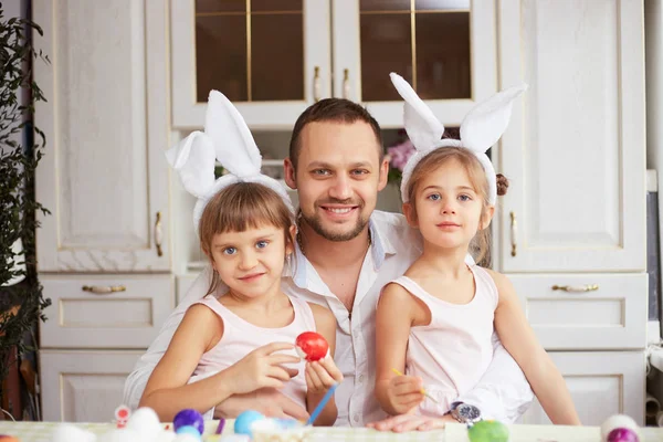 Boldog apa és a két kis lánya a fejükön fehér nyúl füle festék a tojás a húsvét tábla a hangulatos fény konyhában — Stock Fotó