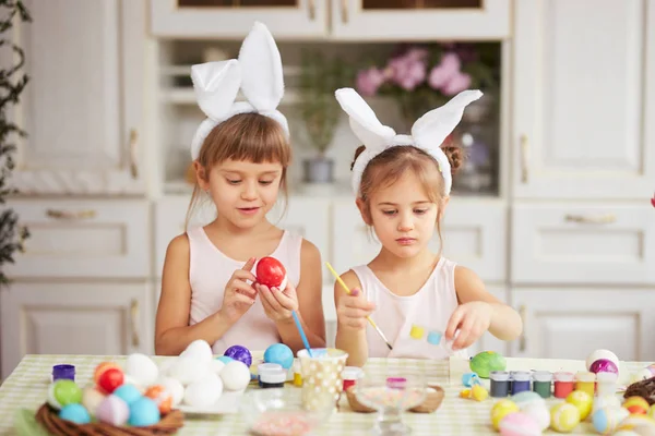 Две маленькие сестры с белыми кроличьими ушами на головах красят яйца для пасхального стола в уютной светлой кухне — стоковое фото