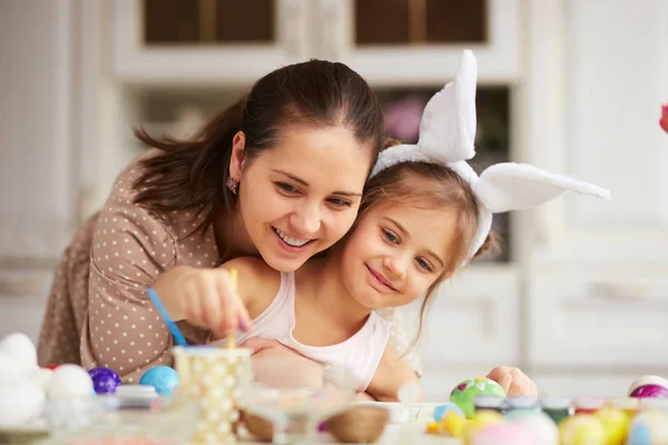 Fiatal anya ölelést vele a kis lányom fehér nyúl füle a fején a tojás a húsvét tábla a hangulatos világos konyha festése — Stock Fotó