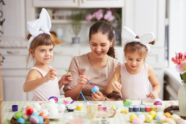 Glückliche junge Mutter und ihre beiden kleinen Töchter mit weißen Hasenohren auf dem Kopf färben die Eier für den Ostertisch in der gemütlich hellen Küche — Stockfoto