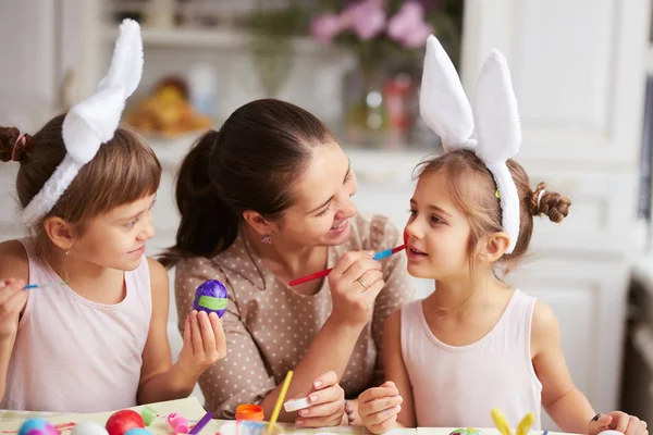 Mutter streicht Töchtern über die Wange, während sie und ihre beiden kleinen Töchter mit weißen Hasenohren auf dem Kopf die Eier für den Ostertisch in der gemütlichen hellen Küche färben. — Stockfoto