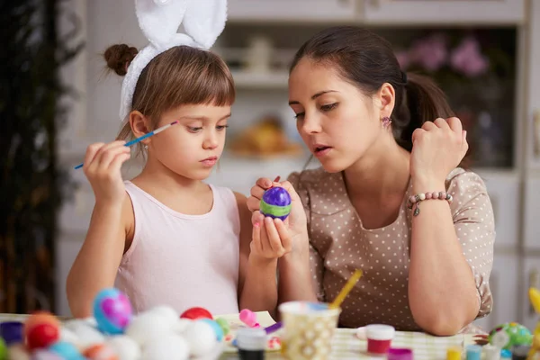 Boldog, fiatal anya és a lány két kis lánya fehér nyúl füle a fejükre festék a tojás a húsvét tábla a hangulatos fény konyhában — Stock Fotó