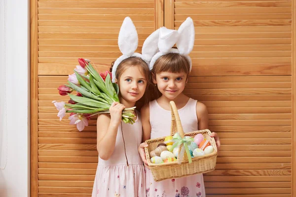 Две очаровательные маленькие сестры в платьях с белыми кроличьими ушами на головах держат цветы и корзину с крашеными яйцами на фоне деревянных дверей — стоковое фото