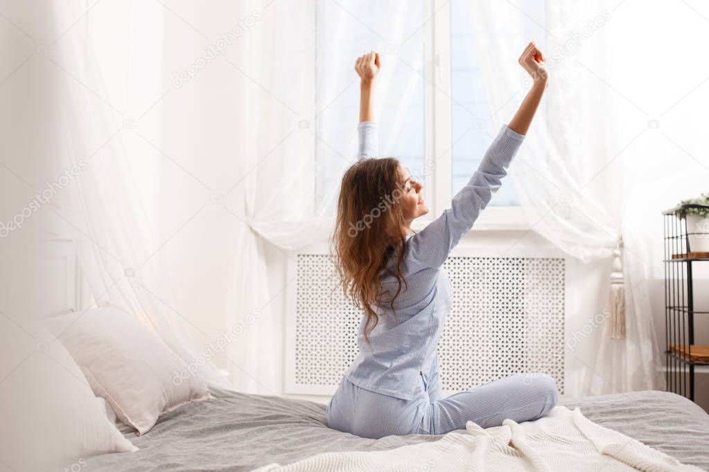 Brunette girl in the light-blue pajama stretches her arms up sitting on the canopy bed next to the window in the cozy bedroom