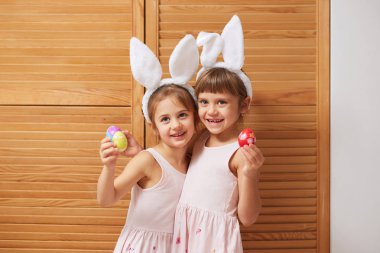 İki komik büyüleyici küçük kız başlarına beyaz tavşan kulaklı elbiseler ellerindeki ahşap kapılar arka plan üzerine boyalı yumurta tutar