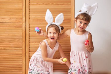 İki komik küçük kız başlarına beyaz tavşan kulaklı elbiseler boyalı yumurta ahşap kapılar arka plan üzerinde ellerinde eğlenin