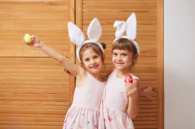 İki komik büyüleyici küçük kız başlarına beyaz tavşan kulaklı elbiseler ellerindeki ahşap kapılar arka plan üzerine boyalı yumurta tutar