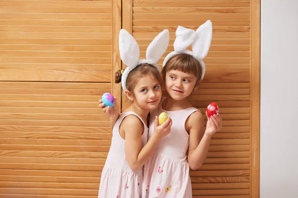 Две забавные очаровательные маленькие сестры в платьях с белыми кроличьими ушами на голове держат окрашенные яйца в руках на фоне деревянных дверей — стоковое фото