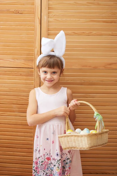 Charmantes kleines Mädchen im Kleid mit weißen Hasenohren auf dem Kopf hält einen Korb mit den gefärbten Eiern auf dem Hintergrund von Holztüren — Stockfoto