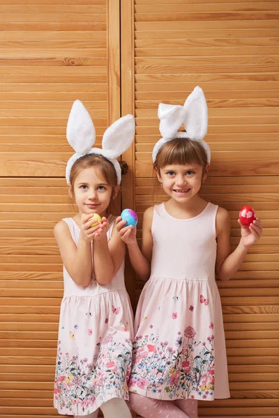 Zwei bezaubernde kleine Schwestern in den Kleidern mit weißen Hasenohren auf dem Kopf halten gefärbte Eier in ihren Händen auf dem Hintergrund von Holztüren — Stockfoto