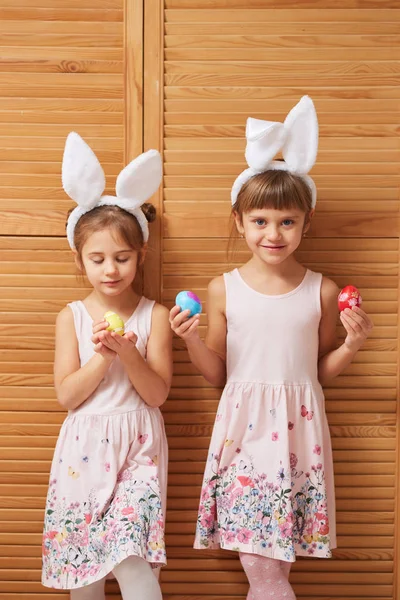 Zwei bezaubernde kleine Schwestern in den Kleidern mit weißen Hasenohren auf dem Kopf halten gefärbte Eier in ihren Händen auf dem Hintergrund von Holztüren — Stockfoto