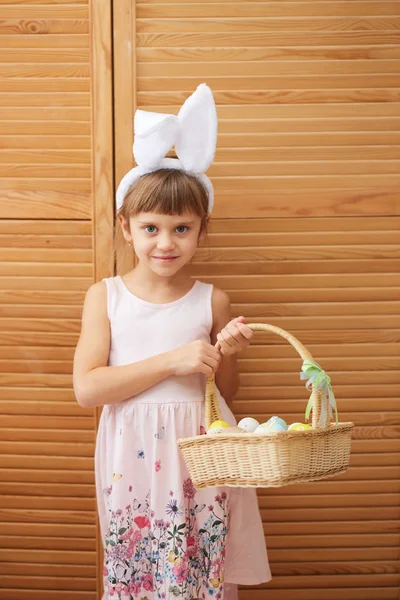 Charmantes kleines Mädchen im Kleid mit weißen Hasenohren auf dem Kopf hält einen Korb mit den gefärbten Eiern auf dem Hintergrund von Holztüren — Stockfoto