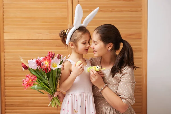 Fiatal gyönyörű anya és vele a kis lányom a ruhában, fején fehér nyúl füle tartani virágok és festett tojásokat a kezükben, a háttérben a fából készült ajtók — Stock Fotó
