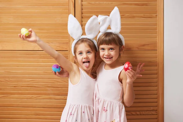 Zwei lustige, charmante kleine Schwestern in den Kleidern mit weißen Hasenohren auf dem Kopf halten gefärbte Eier in ihren Händen auf dem Hintergrund von Holztüren — Stockfoto