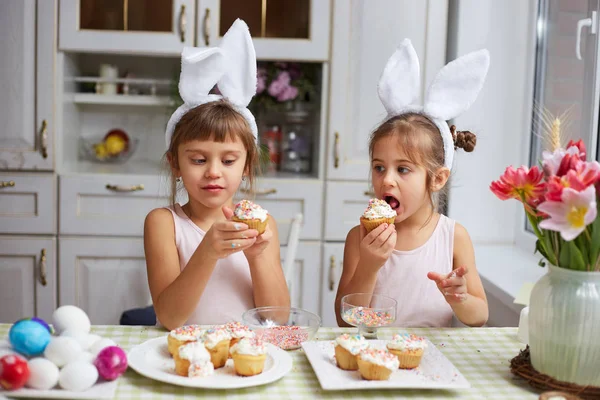 Две маленькие сестры с белыми кроличьими ушами на голове едят маленькие пасхальные торты на уютной светлой кухне. — стоковое фото