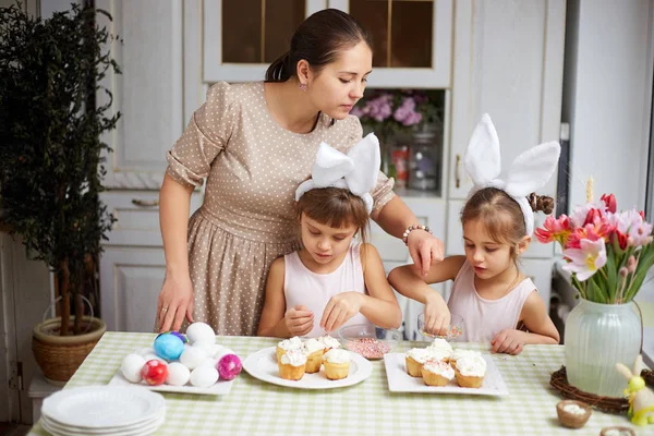 Ung mamma och hennes två små döttrar med vit kanin öron på huvudet laga små påsk kakor för tabellen påsk i den mysiga ljus kök — Stockfoto