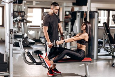 Siyah spor kıyafetlerini giymiş güzel ince kız spor salonunda antrenör gözetiminde özel egzersiz makinede karın için egzersizler yapıyor