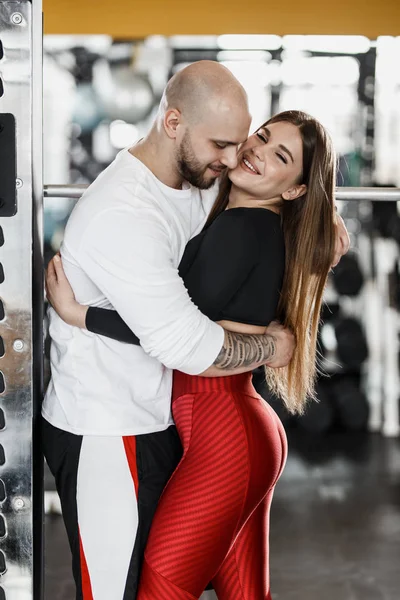 浪漫快乐运动的夫妇。强壮的男人和苗条美丽的女孩拥抱在运动器材旁边的现代健身房里 — 图库照片