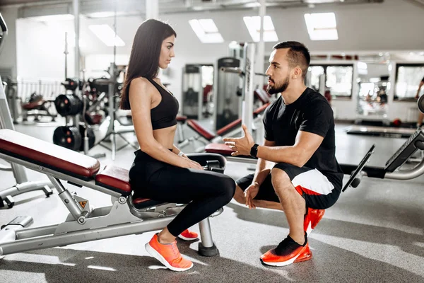 Coach talks with athletic girl dressed in black sport clothes sitting on the bench in the gym