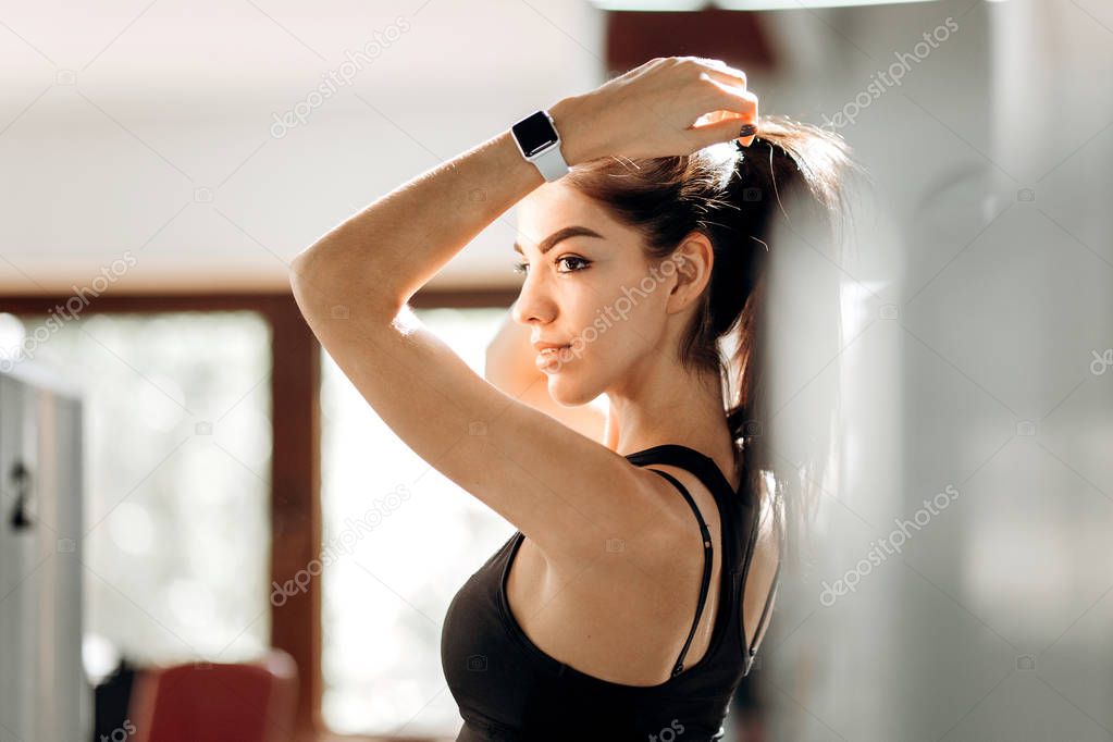 Beautiful slim girl dressed in black sportswear is standing in the gym and tying her hair