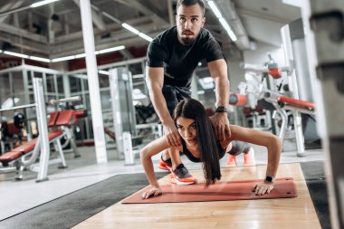 Siyah spor top ve tayt giymiş güzel atletik kız tahta yapıyor ve Koç onu spor salonunda düzeltir