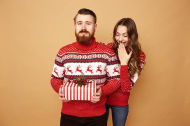 Guy dressed in red and white sweater with deer holds a Christmas present in his hands and a girl looks out from behind his back on a beige background in the studio clipart