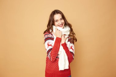 Beautiful girl dressed in a red and white sweater with deer and white knitted scarf stands on a beige background in the studio clipart