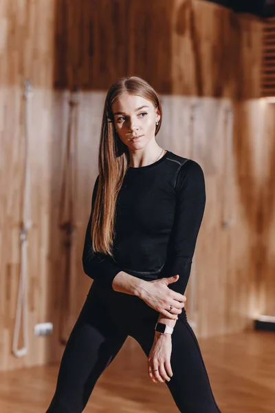 Красива струнка дівчина, одягнена в чорний спортивний одяг стоїть в сучасному тренажерному залі з дерев'яними прикрасами — стокове фото