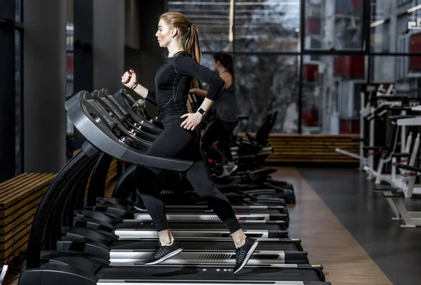 La chica atlética vestida con una ropa deportiva negra corriendo en la cinta en el moderno gimnasio — Foto de Stock