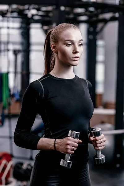 Стройная молодая девушка в черной спортивной одежде делает упражнения с гантелями в современном тренажерном зале с большим количеством спортивного оборудования — стоковое фото