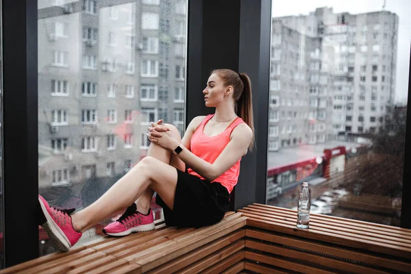 The athletic girl dressed sport shorts and t-shirt is sitting on the wooden windowsill in the modern gym