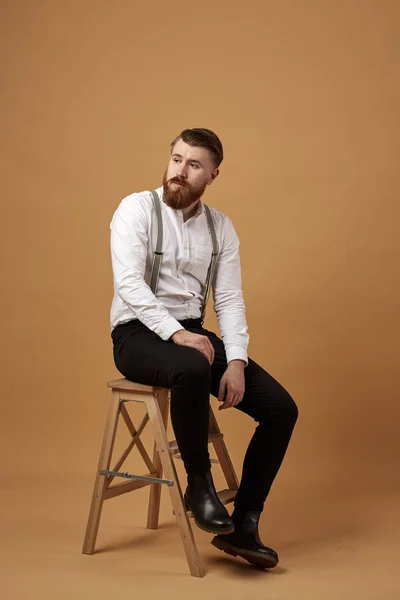 Stijlvolle roodharige man met baard, gekleed in een wit overhemd en een zwarte broek met jarretellen zit op een houten kruk op een beige achtergrond — Stockfoto