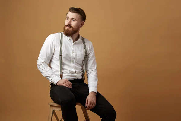 Elegante hombre pelirrojo con barba vestida con una camisa blanca y pantalones negros con tirante sentado en un taburete de madera sobre un fondo beige — Foto de Stock