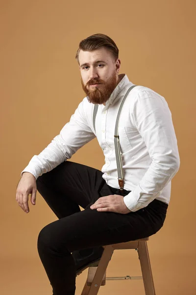 Stylish red-haired man with beard dressed in a white shirt and black trousers with suspender sits on a wooden stool on a beige background — Stock Photo, Image