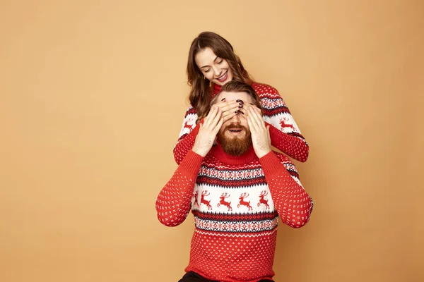 Girl closes the guys eyes with her hands on a beige background in the studio.They both dressed in red and white sweaters with deer. — Stock Photo, Image