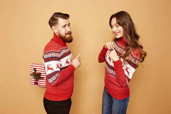 Red-haired guy gives a Christmas gift to a girl both are dressed in red and white sweaters with deer and stands on a beige background in the studio