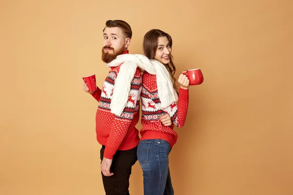 Дівчина і хлопець, одягнені в червоні та білі светри з оленями та білим в'язаним шарфом, стоять на спині і тримають червоні чашки на бежевому тлі — стокове фото