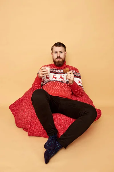 Рудий чоловік з бородою, одягнений в червоно-білий светр з оленями і червоним мисом сидить на підлозі і тримає склянку молока і печива на бежевому фоні — стокове фото