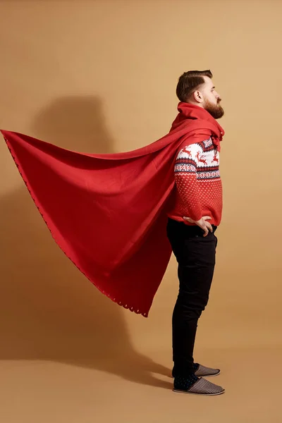Rothaarige Mann mit Bart in einem rot-weißen Pullover mit Hirsch und rotem Umhang flattert im Wind und Schläfer steht auf einem beigen Hintergrund — Stockfoto
