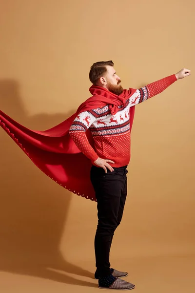 Homem de cabelos vermelhos com barba vestida com uma camisola vermelha e branca com veados e capa vermelha batendo com o vento e travessas fica em um fundo bege — Fotografia de Stock