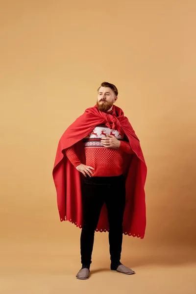 Homme aux cheveux roux avec barbe vêtu d'un pull rouge et blanc avec cerf et cape rouge et traverses se tient sur un fond beige — Photo