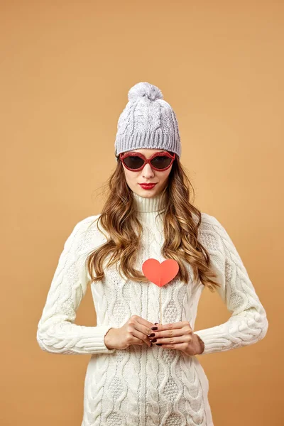 Chica divertida en gafas de sol vestida con suéter de punto blanco y sombrero se divierte con un corazón de papel rojo en un palo sobre un fondo beige en el estudio — Foto de Stock