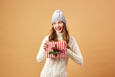 Beyaz örgü Kazak giymiş kız gülümseyerek ve stüdyoda bej renkli bir arka plan üzerinde bir Noel hediyesi şapka tutar