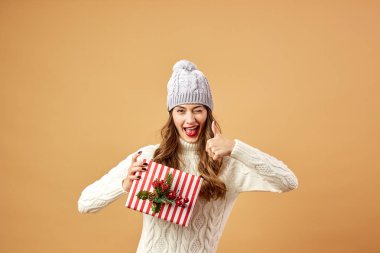 Beyaz örgü Kazak giymiş kız gülümseyerek ve stüdyoda bej renkli bir arka plan üzerinde bir Noel hediyesi şapka tutar