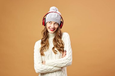 Kırmızı Kulaklığımda güzel kız beyaz örgü Kazak giymiş ve şapka stüdyoda bej renkli bir arka plan üzerinde duruyor