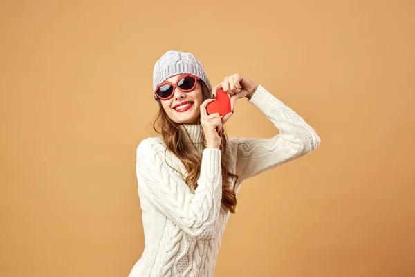 Lustiges Mädchen mit Sonnenbrille, weißem Strickpullover und Hut amüsiert sich mit einem roten Papierherz auf einem Stock auf beigem Hintergrund im Studio — Stockfoto