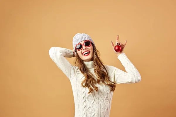 Lächelndes Mädchen mit Sonnenbrille, weißem Strickpullover und Hut hält im Studio eine rote Weihnachtskugel auf beigem Hintergrund — Stockfoto